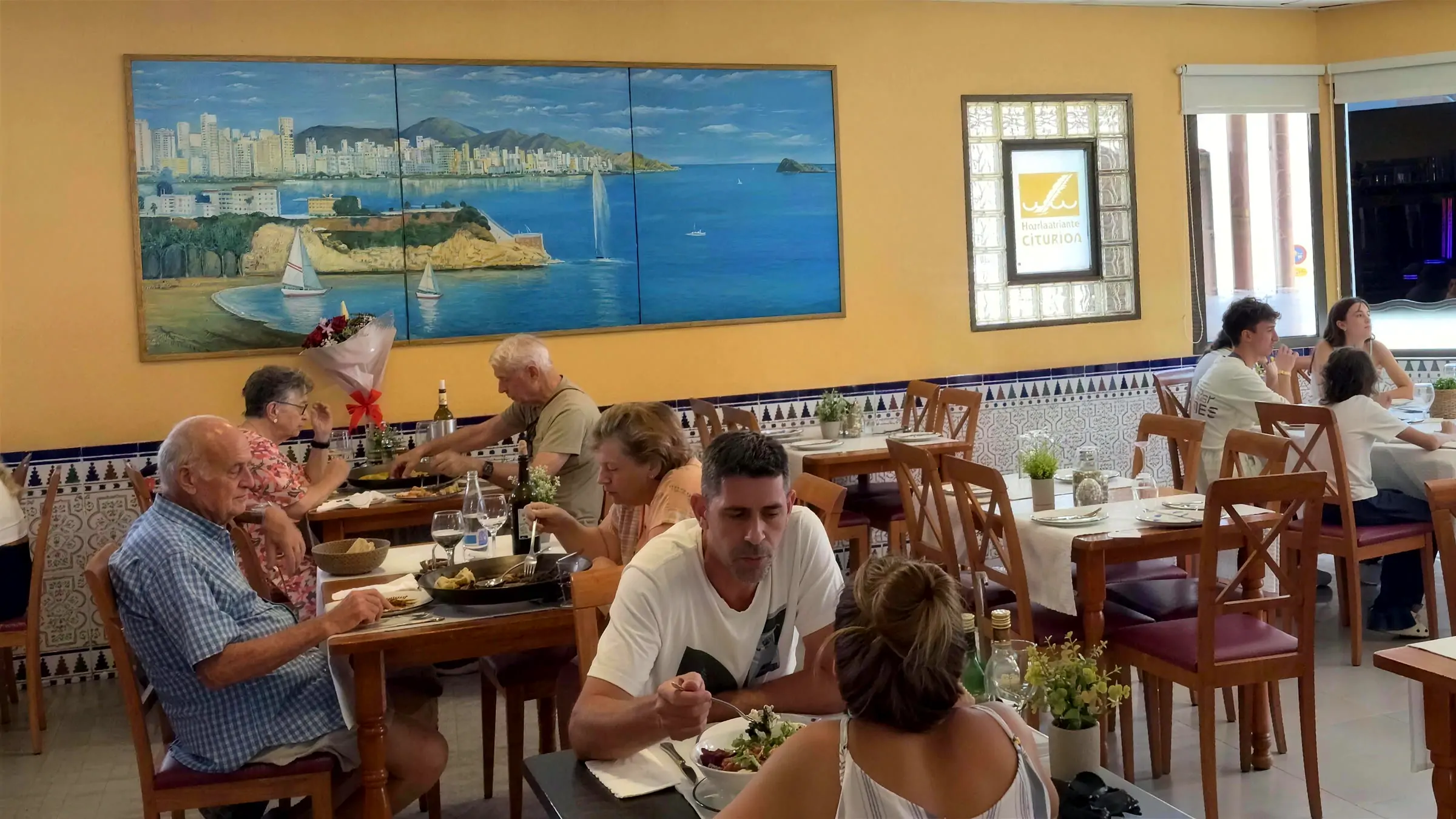 The Esturión dining room with guests having lunch and the mural of Benidorm bay